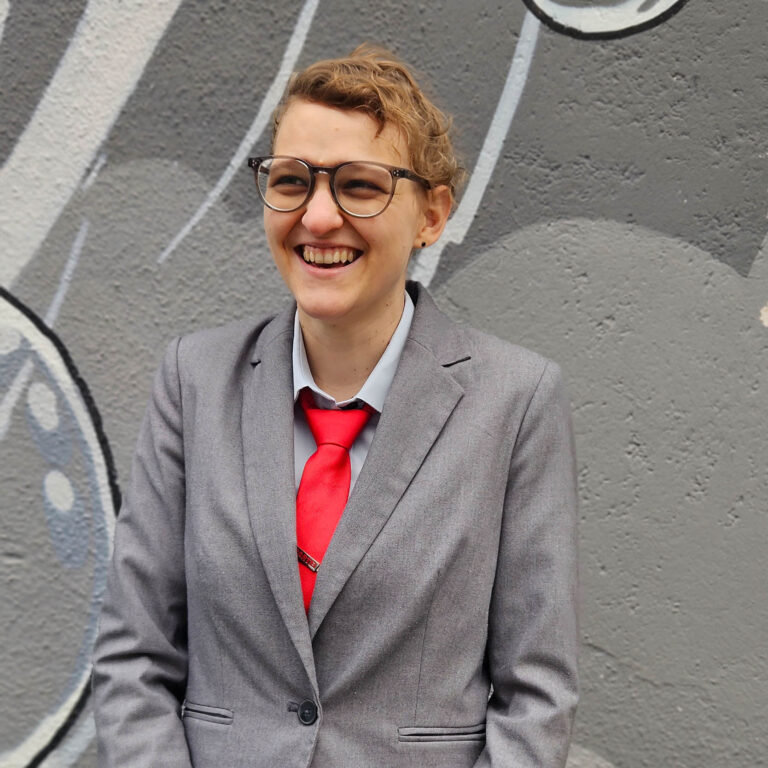 KLara Schinnerl, dunkeblonde kurze lockige Haare, Brille, steht in grauem Anzug mit roter Krawatte fr&ouml;hlich lachend vor einer mit grauen Farben bespr&uuml;hten Wand